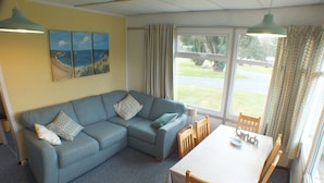A living room with a blue sofa, a dining table with chairs, and a painting of a beach scene on the wall. -  Chalet Cwtch, a cosy 5 bed holiday cottage in Roch, Pembrokeshire (Roch)
