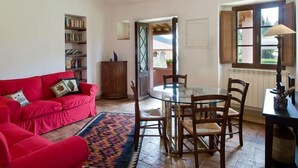 A cozy living room with a red sofa, a glass dining table with chairs, a bookshelf, and a view through a door to an outdoor area. - Montestigliano, an authentic farmhouse in the heart of Tuscany, is an oasis of peace and beauty that offers its guests refined residences and an enchanting view (Rosia - Sovicille)