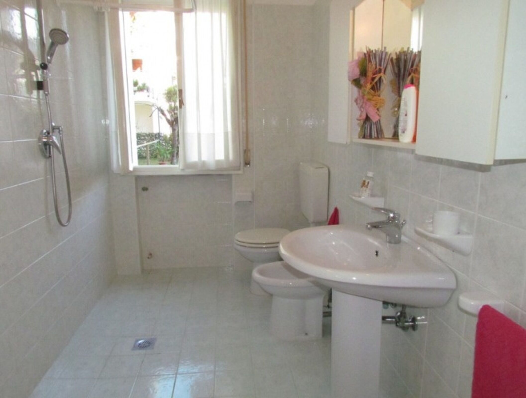 Ein Badezimmer mit Dusche, Toilette und Waschbecken, mit weißen Fliesen und einem Fenster mit Vorhängen.