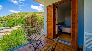 A balcony with a wooden table and bench, a view of a green hillside, and a room with a bed and a desk. - Holiday home on the amazing Cilento Coast in the south of Italy (Case del Conte)