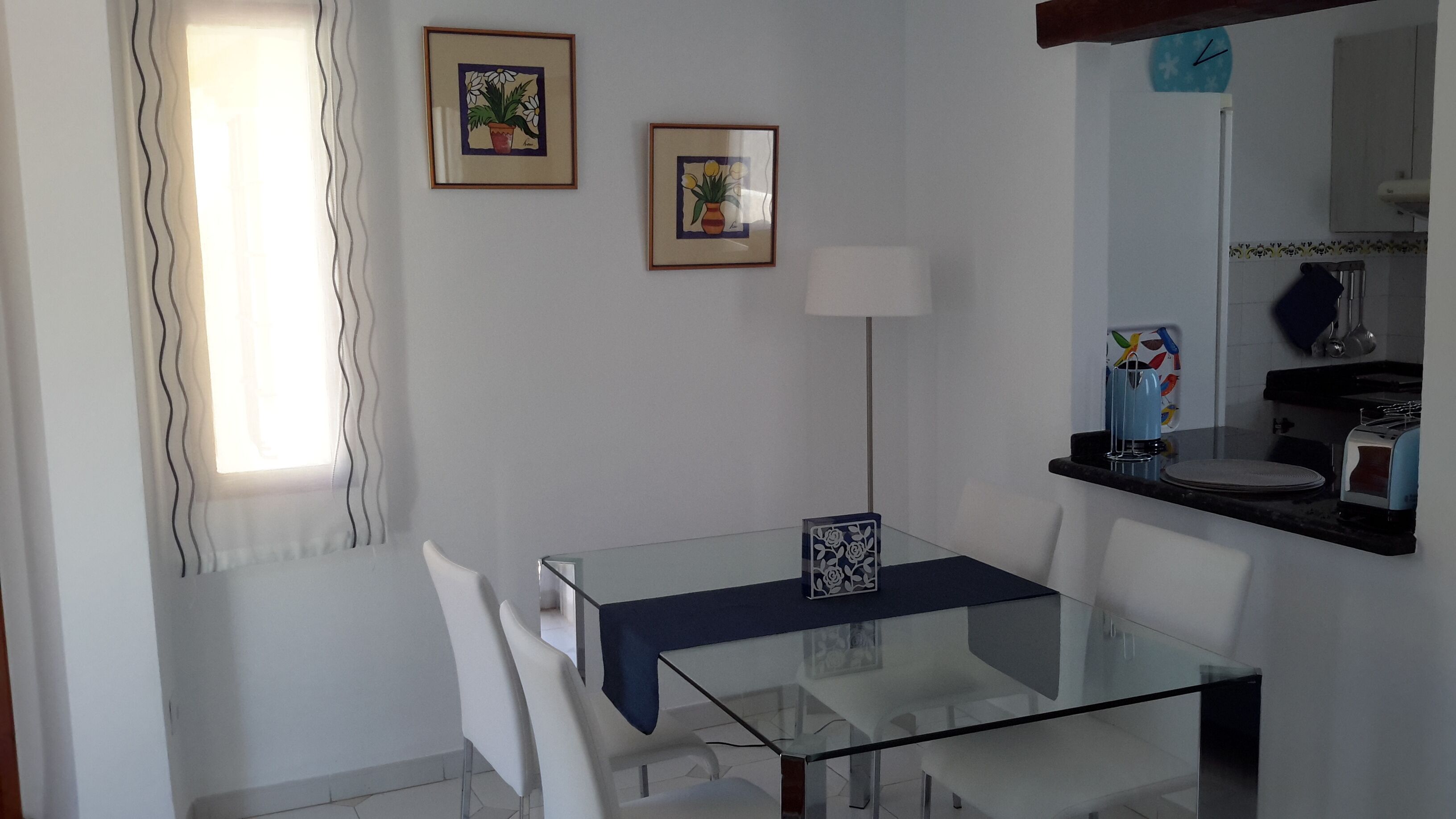 Un coin repas avec une table en verre, des chaises blanches, une nappe bleue et deux œuvres d’art encadrées accrochées au mur.