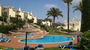 A pool area with blue lounge chairs, palm trees, and buildings with Mediterranean architecture. - Inviting, bright 2-Bedroom El Rancho villa in prestigious La Manga Club Resort (La Manga Club)