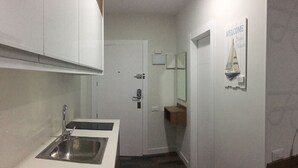 Un couloir étroit, avec un coin cuisine sur la gauche, une porte ornée d’un miroir et d’une pancarte de bienvenue sur la droite, et une petite armoire en bois située sous la porte.