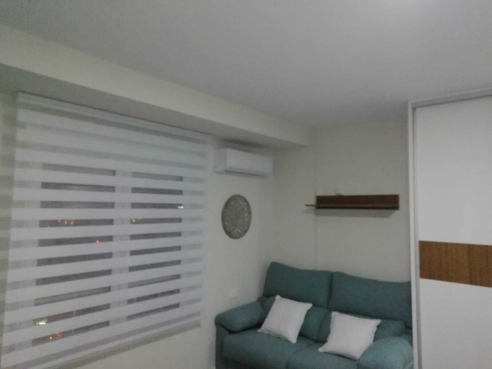 Una habitación con ventana con persianas horizontales, un sofá azul verdoso con cojines blancos, un estante de madera y un adorno redondo en la pared.