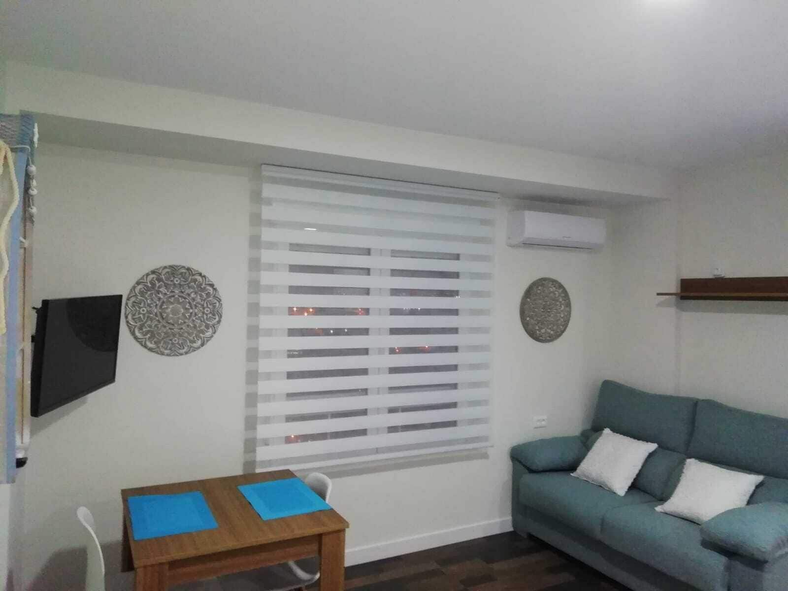 Una sala de estar con un sofá, una mesa con individuales azules, un televisor, un ventanal con persianas y decoración en la pared.