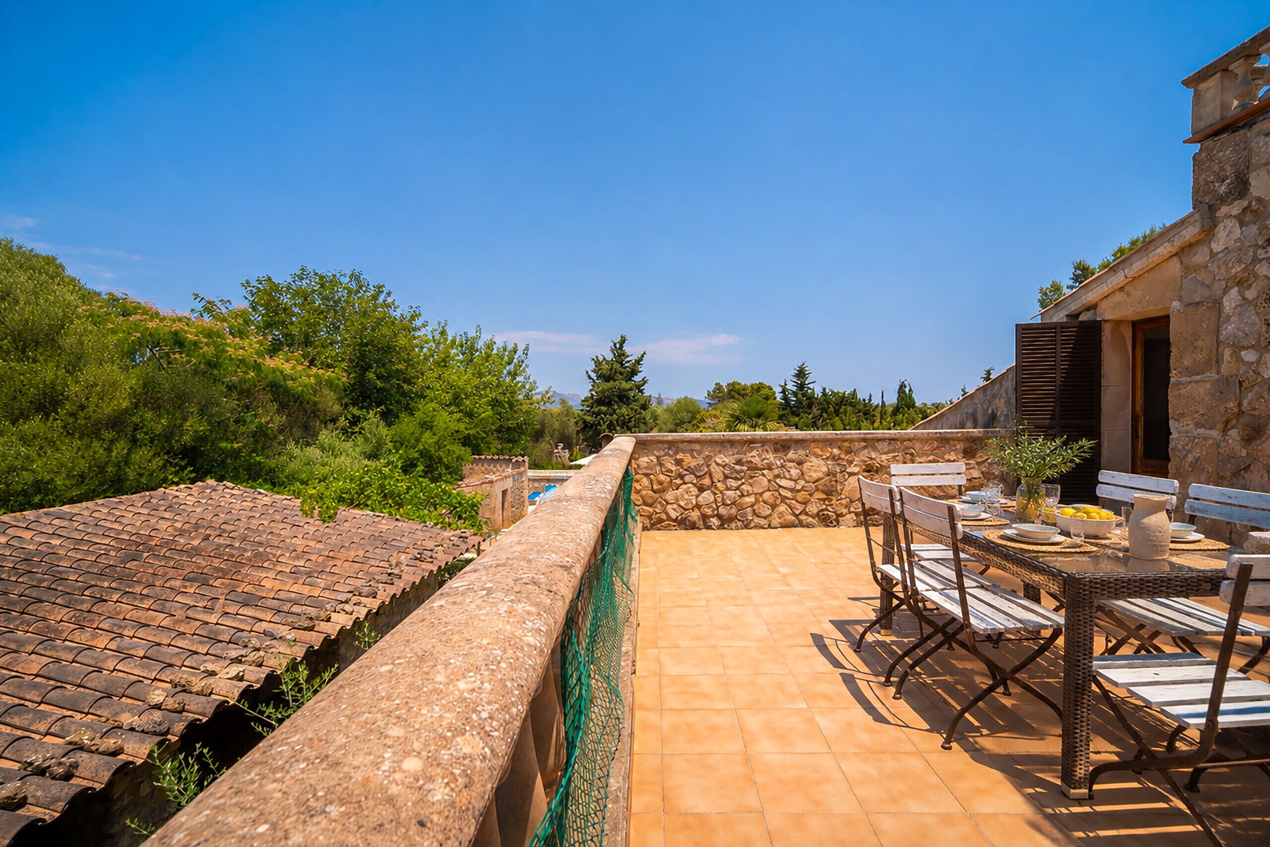 A rooftop terrace with a stone wall, a table with chairs, and a view of greenery and distant mountains.