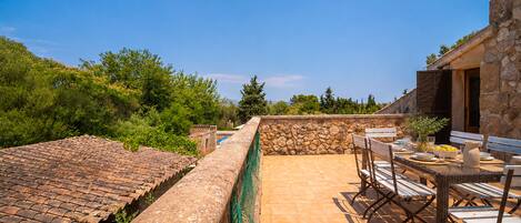 A rooftop terrace with a stone wall, a table with chairs, and a view of greenery and distant mountains.