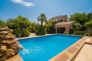 A swimming pool with a stone border, surrounded by greenery and a stone building with a tiled roof.