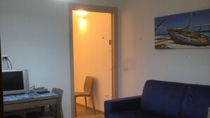 A room with a blue sofa, a desk with a computer, a chair, and a painting of a boat on the wall. - Two-room apartment in Palau, 20 minutes from the Costa Smeralda in Sardinia (PALAU (OT))