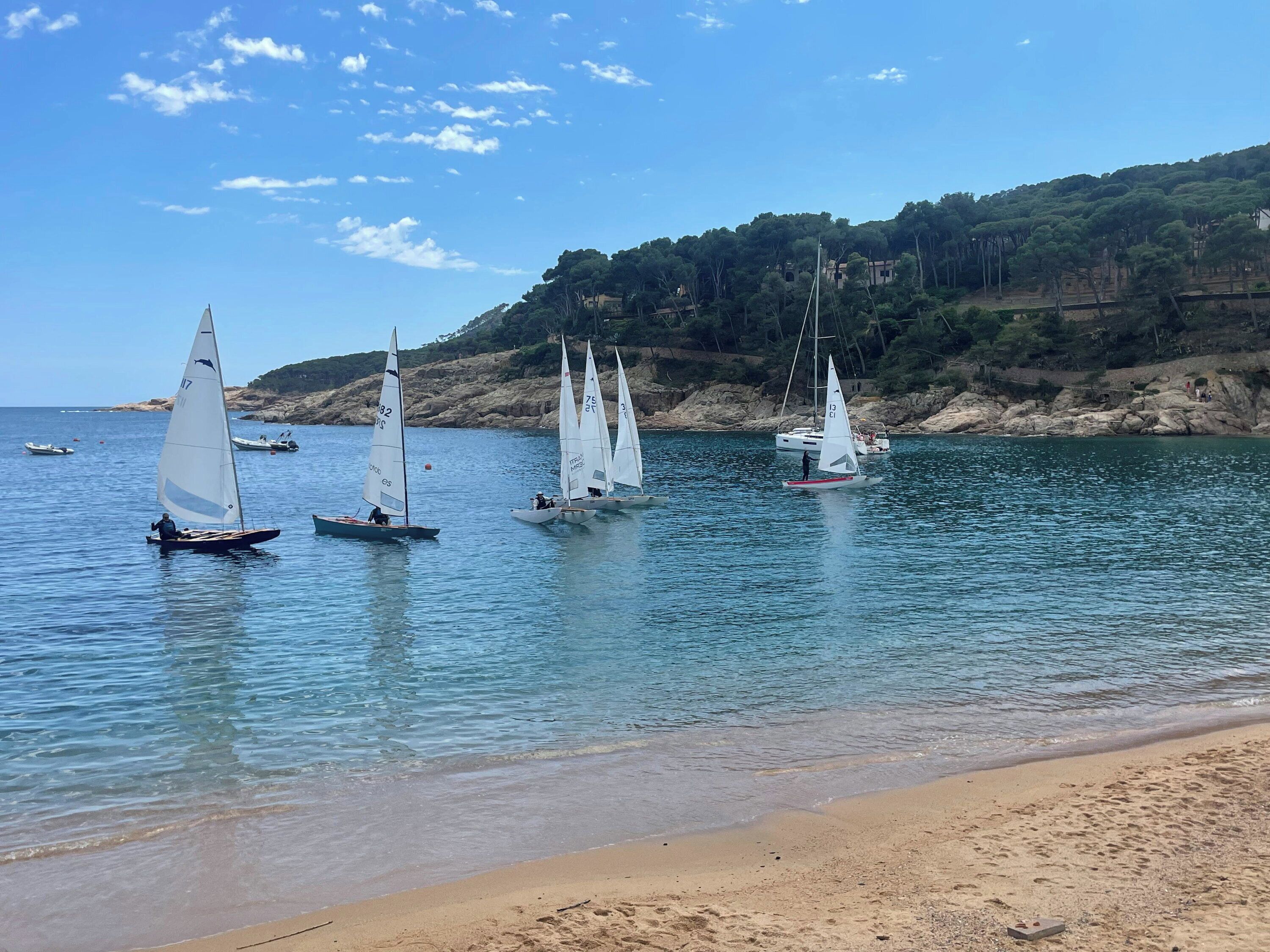 A beach with sailboats on calm waters, a sandy shore, and a hilly backdrop.