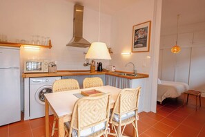 A compact kitchen with a washing machine, dining table, wicker chairs, and a bed in the adjacent room. - ApartmentsTamariu#5 🌊 Cozy Retreat with Terrace (Tamariu)