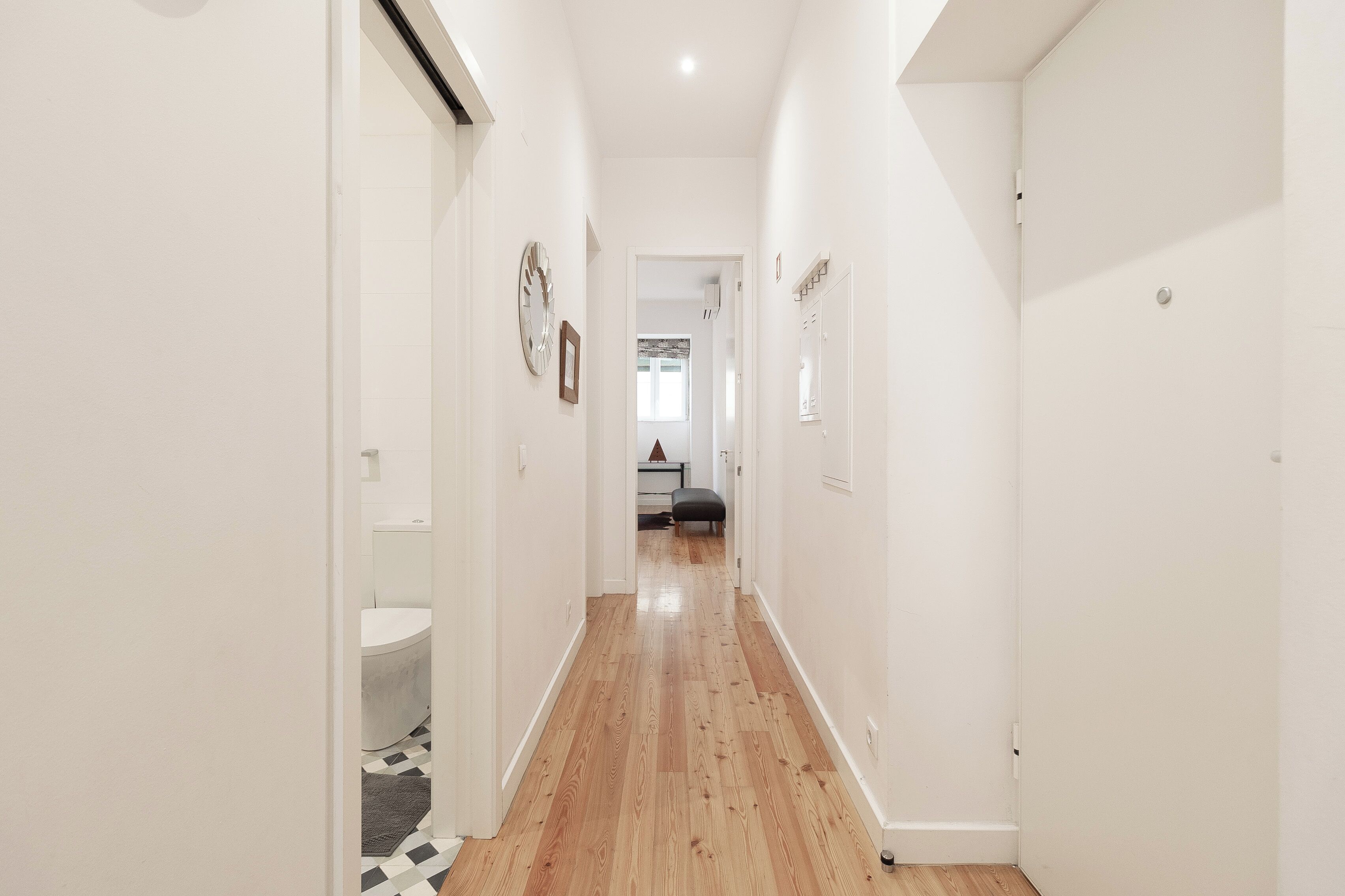 Un pasillo estrecho con suelo de madera y paredes blancas que conduce a un baño con inodoro y lavamanos.