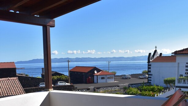 Ein Balkon mit roten Stühlen und einem Blumentopf, mit Blick auf eine Küstenlandschaft mit Gebäuden und blauem Himmel.
