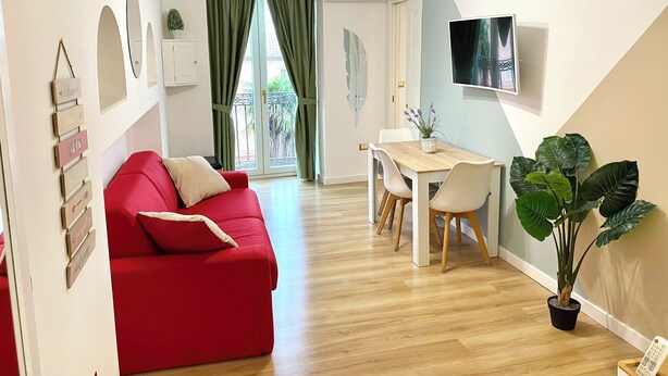A modern living room with a red sofa, a dining area with a wooden table and chairs, a TV mounted on the wall, and a large window with green curtains. - SuiteCorso Vibo Valentia (Vibo Valentia)