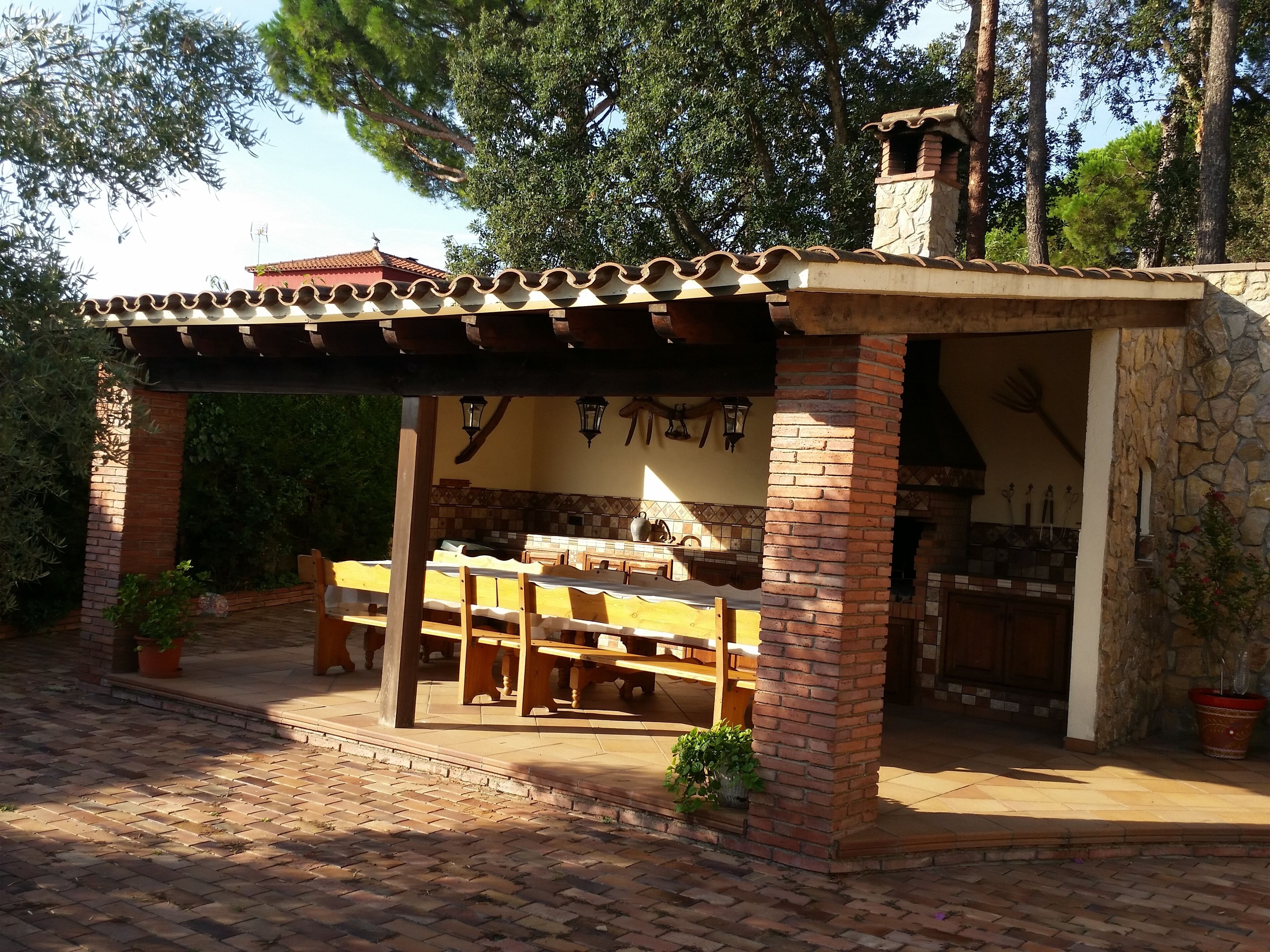 A covered outdoor dining area with brick walls and a tiled roof, featuring wooden benches and tables.