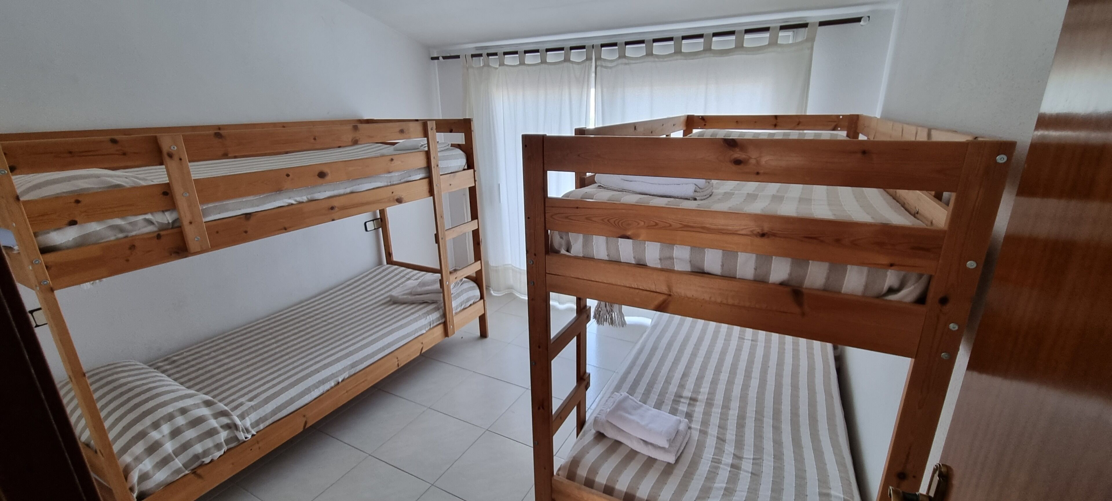 A room with two bunk beds, a window with curtains, and a white tiled floor.