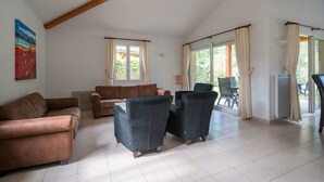 Amplia sala de estar con un sofá de cuero marrón, dos sillones oscuros y una mesa de centro. Hay ventanas grandes con cortinas, un cuadro en la pared y vistas al exterior.