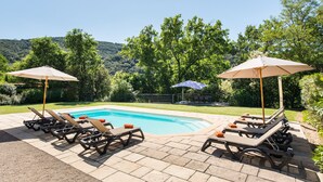 Un área junto a la piscina con sillones de descanso y sombrillas, rodeada de abundante vegetación y colinas.