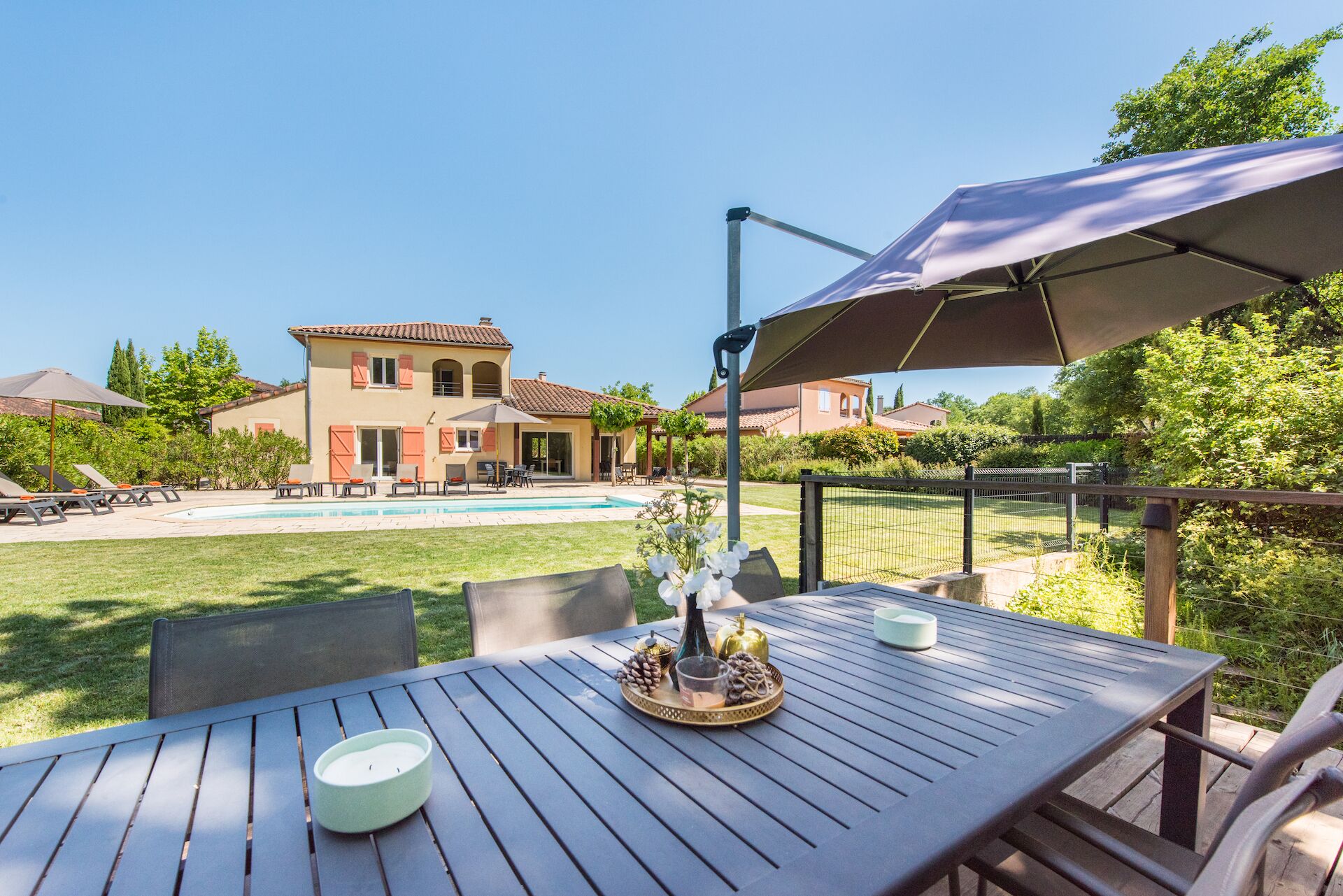 Un patio con una mesa lista para comer, una piscina y una casa al fondo.