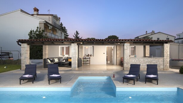 Ein Wohnhaus mit Schwimmbad, Liegestühlen, einer überdachten Terrasse und einer Fassade aus Naturstein.