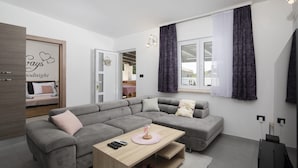 Ein modernes Wohnzimmer mit einem grauen Ecksofa, einem Holztisch und Blick auf einen Balkon durch ein Fenster mit violetten Vorhängen.