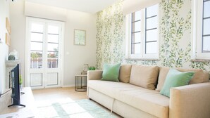Un salon avec un canapé beige, des coussins verts, une petite table d’appoint et un téléviseur. Une fenêtre donne sur un balcon.