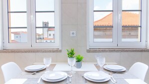 Une table à manger dressée pour quatre personnes, avec de l’argenterie, de la vaisselle blanche et des verres, placée entre deux fenêtres offrant une vue sur les bâtiments.