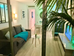 Uma sala de estar moderna com um sofá cinza, manta azul-esverdeada e uma mesa de centro de vidro. Uma televisão está fixada na parede, e um vaso de planta grande está em primeiro plano.