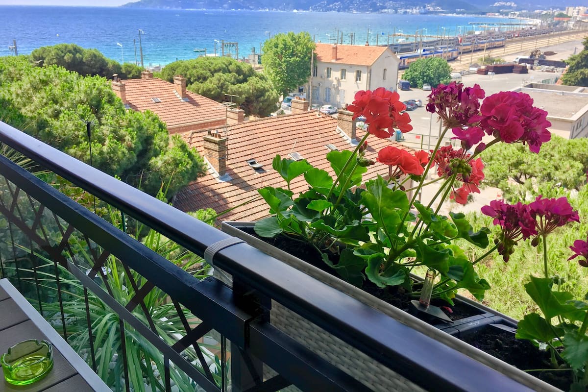 A balcony with potted geraniums, a clear view of the sea, and a coastal town in the background.