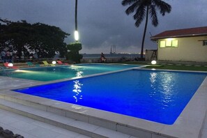 A swimming pool with two sections, surrounded by lounge chairs and palm trees, with a view of a distant building. - Fini Hotel Bobende (Limbe)