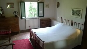 Een slaapkamer met een enkel bed, een dressoir, een stoel, een radiator en een raam met open luiken.