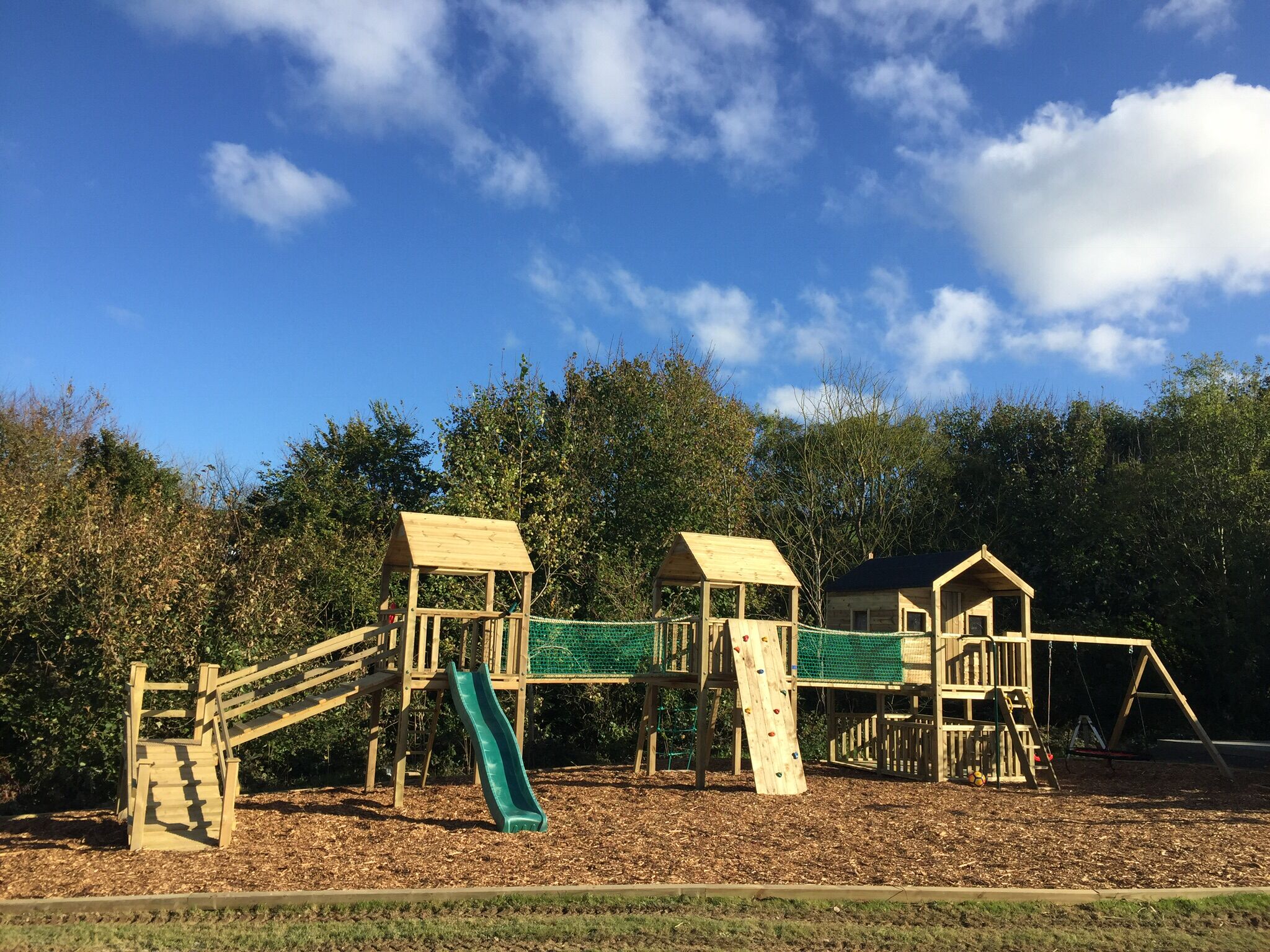 A wooden playground with slides and climbing structures, surrounded by trees and a clear sky.