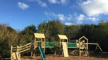 A wooden playground with slides and climbing structures, surrounded by trees and a clear sky.