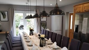 Una larga mesa de comedor dispuesta para una comida, con múltiples sillas, iluminación colgante y vistas al exterior a través de amplios ventanales.