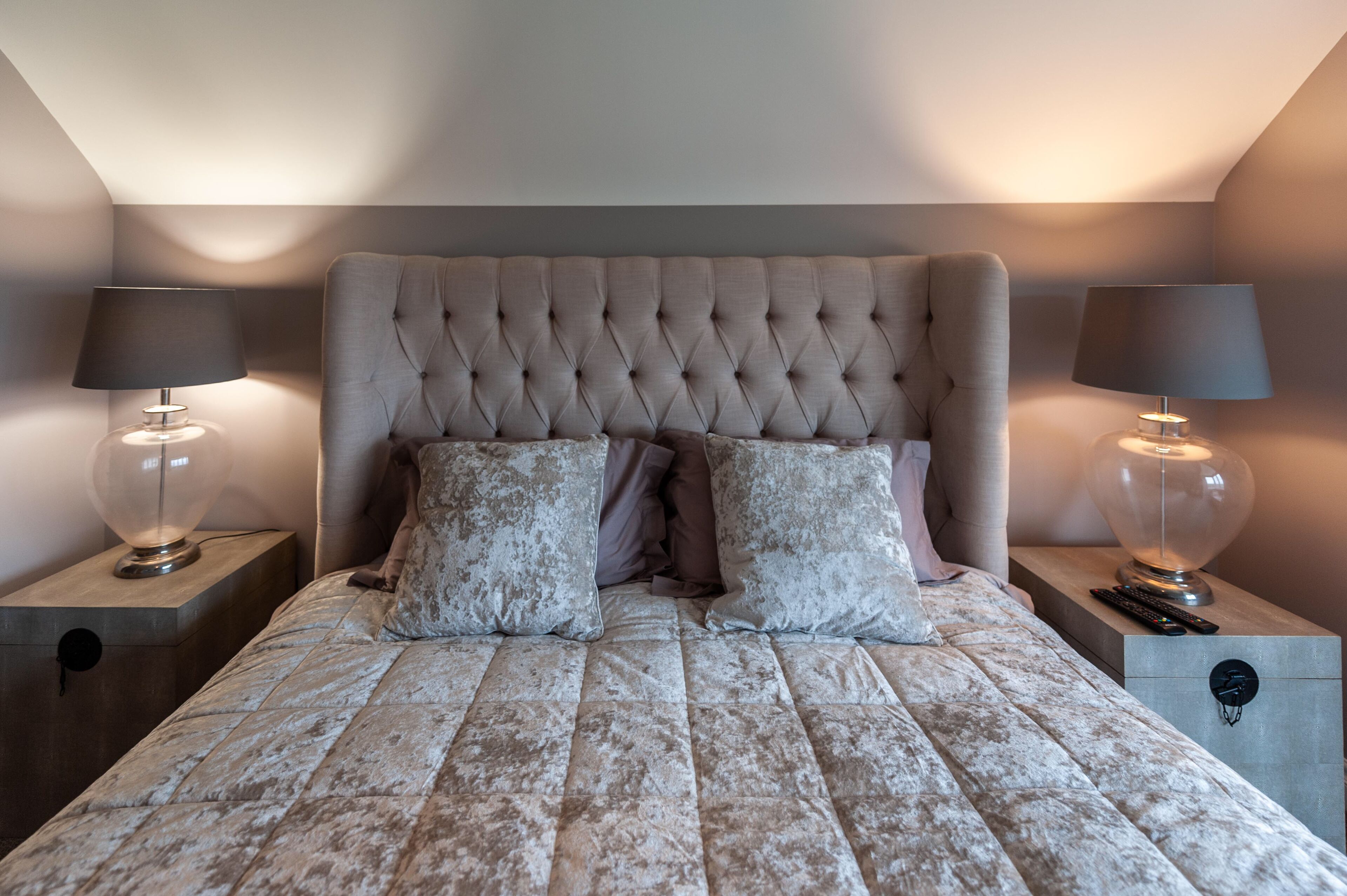 A bedroom with a tufted headboard, two bedside lamps, a bed with patterned bedding, and a nightstand with a remote control.