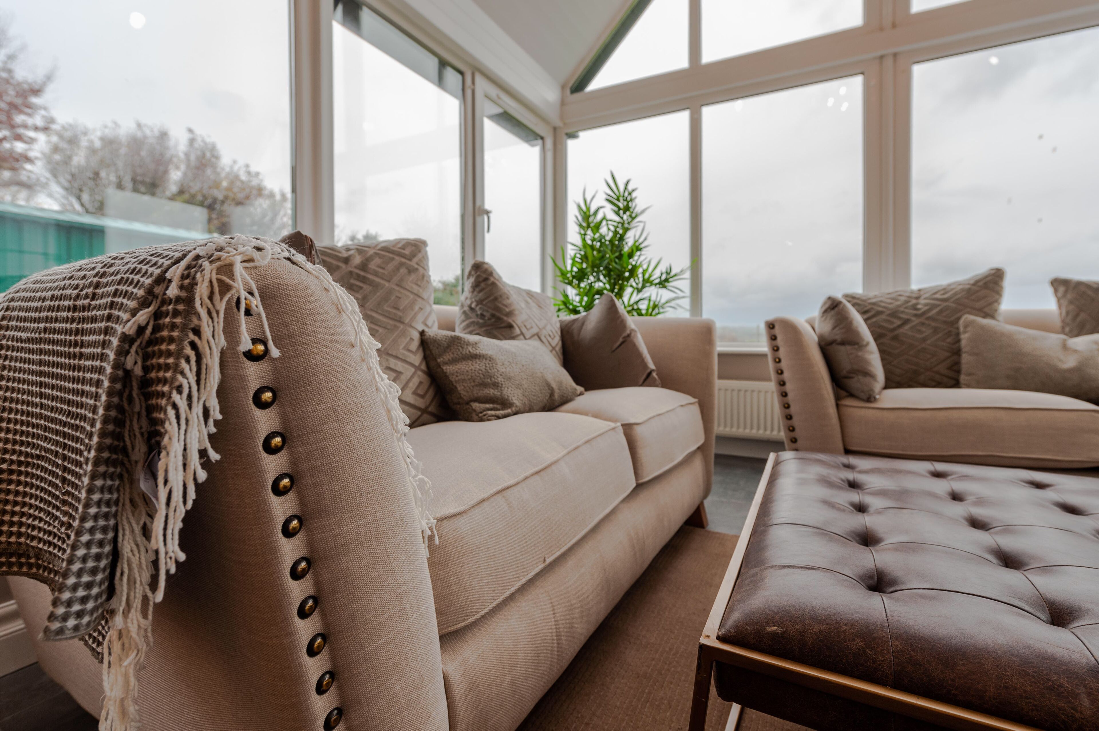 Ein gemütliches Wohnzimmer mit einem beigefarbenen Sofa, einem braunen Hocker und einem großen Fenster mit Blick ins Freie.