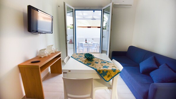 A compact living space with a TV mounted on the wall, a wooden bench, a dining table with a table runner, and a blue sofa.