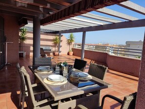 Eine Terrassensitzgruppe im Freien mit Esstisch, umgeben von Korbstühlen und einem überdachten Pergola.