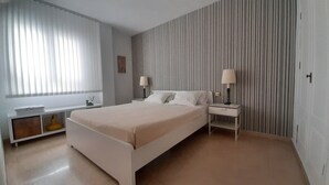 Un dormitorio con cama, mesitas de noche, lámpara, ventana con persianas y pared a rayas verticales.
