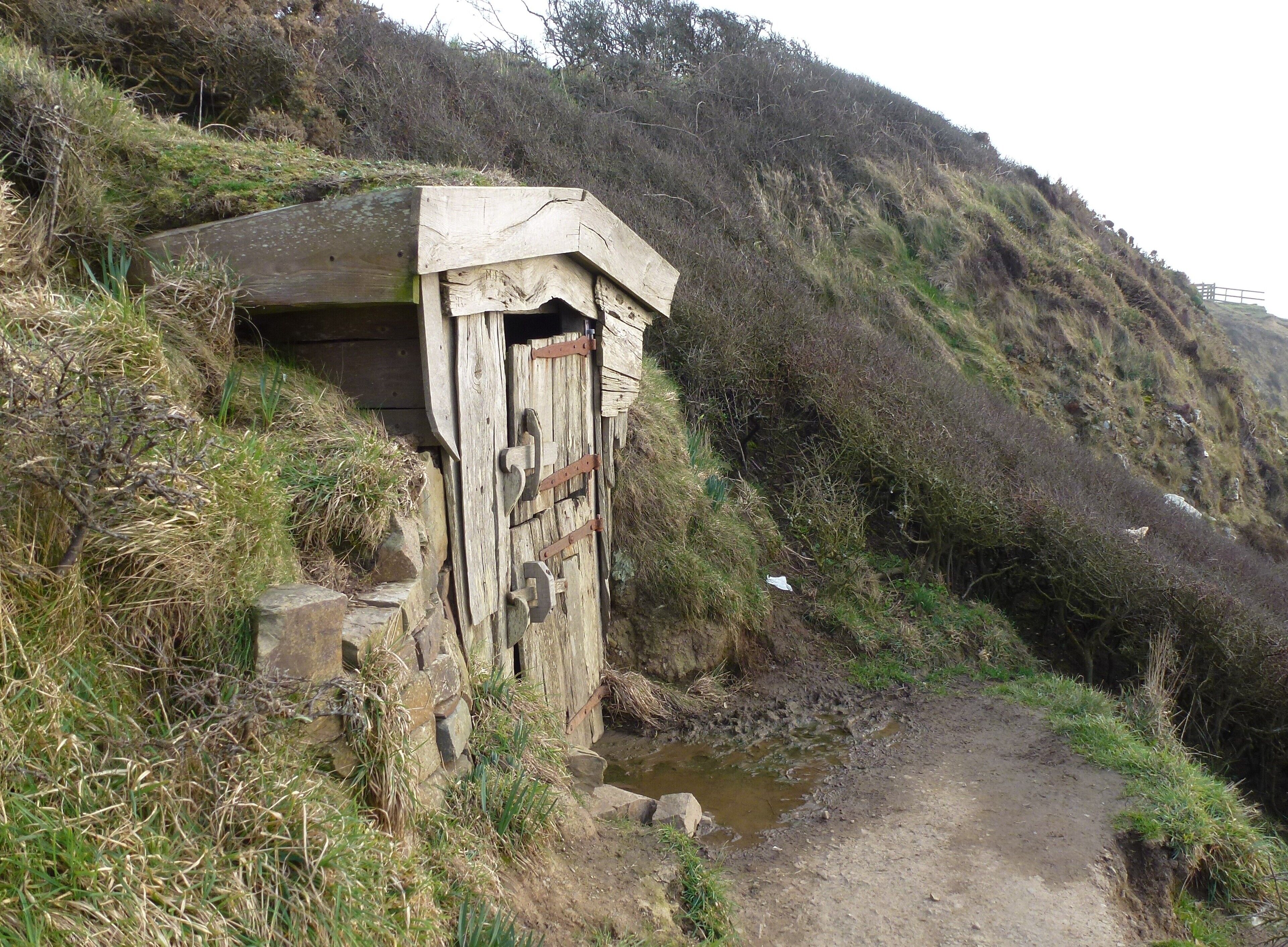 A rustic wooden structure with a door, situated beside a narrow, muddy path.