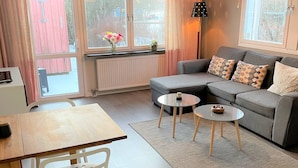 Un moderno salón con un sofá gris, una mesa de comedor de madera y una silla blanca. Hay un ventanal con cortinas, una lámpara colgante y una pequeña alfombra en el suelo.