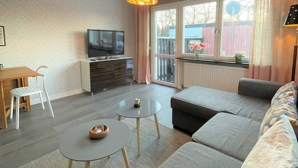 Una sala de estar moderna con sofá, dos mesas redondas, televisor y balcón con vistas.