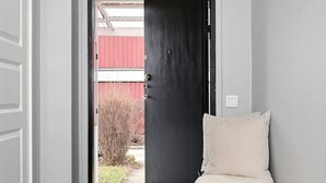 Un pasillo con una puerta negra que da al exterior, un banco blanco con un cubrefrío mullido y un piso de baldosas.