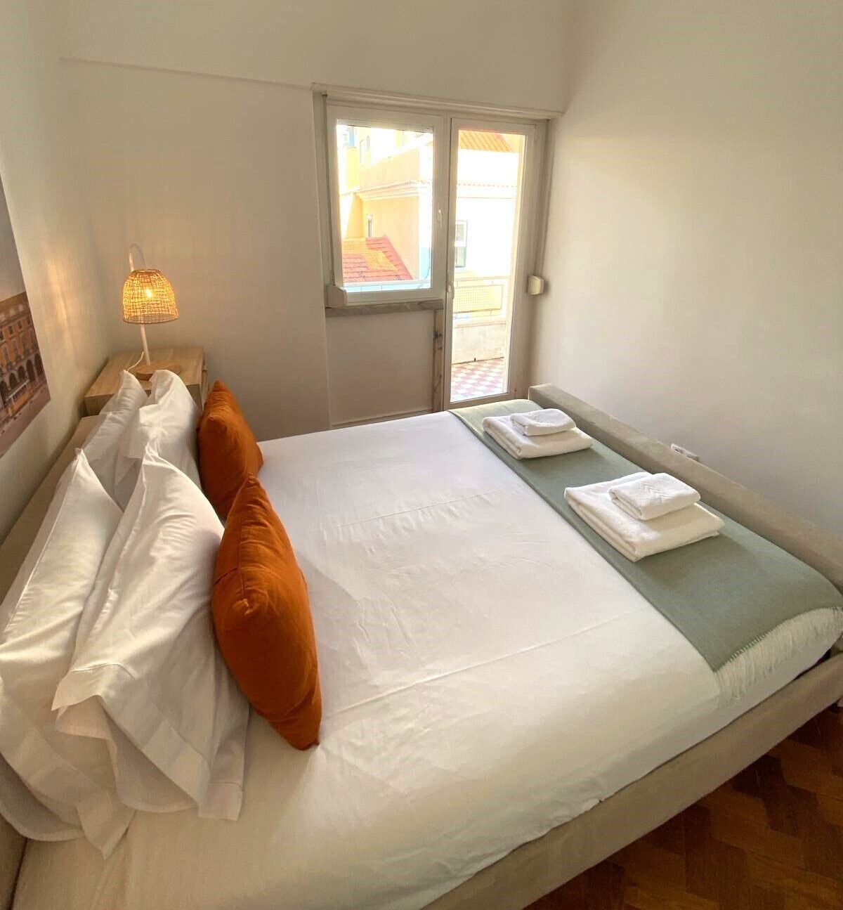 Ein ordentlich bezogenes Bett mit weißen und orangefarbenen Kissen, einer Nachttischlampe und einem Fenster mit Blick auf Gebäude.
