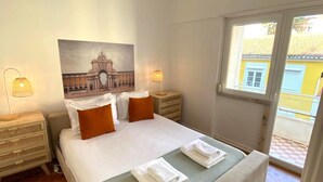 Un cuarto de hotel con una cama grande, dos mesitas de noche, un cuadro de un puente y vista a un edificio amarillo a través de una ventana.