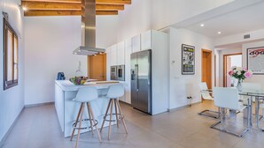 A modern kitchen with white cabinetry, stainless steel appliances, and wooden beams on the ceiling. There are two bar stools and a dining table with chairs. - Villa Can Salat Selva (Selva)