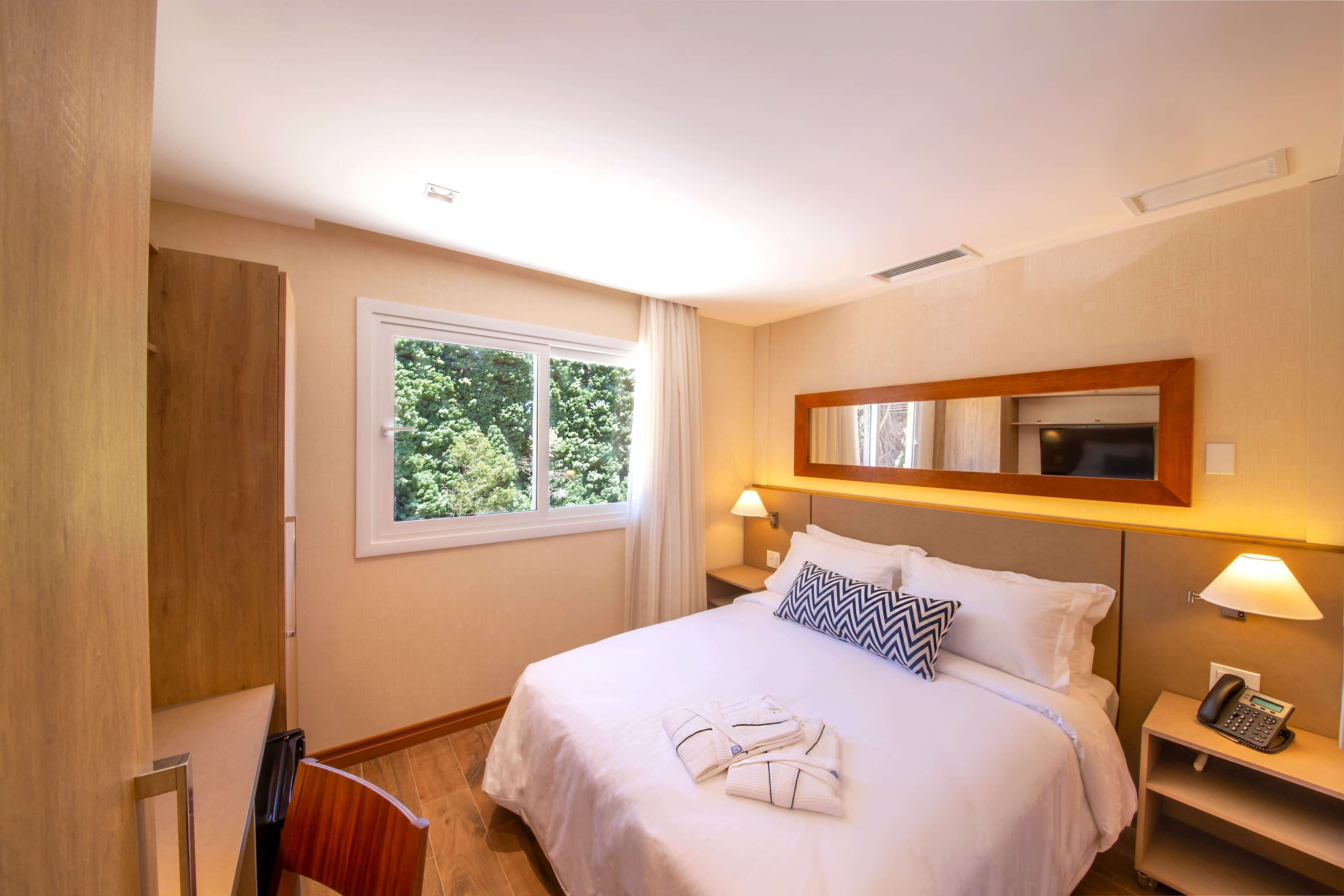 A hotel room with a bed, bedside lamps, a telephone, and a window with a view of trees.