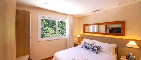 A hotel room with a bed, bedside lamps, a telephone, and a window with a view of trees.