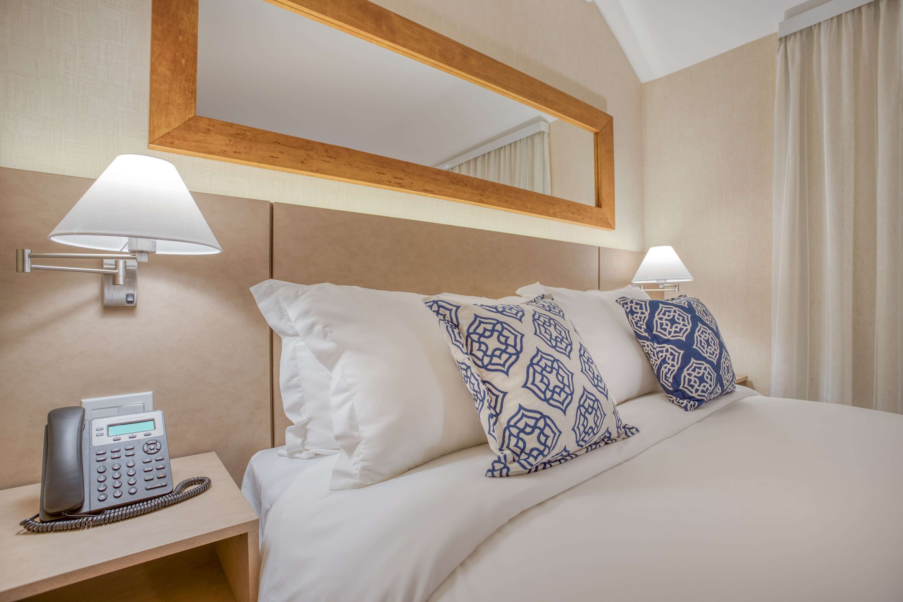 Habitación de hotel con cama, dos almohadas estampadas, lámpara de noche, teléfono y espejo sobre la cama.