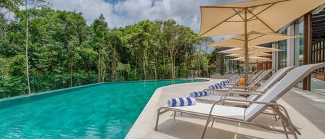 Zona junto a la piscina con sillones y sombrillas, rodeada de árboles y un cielo despejado.